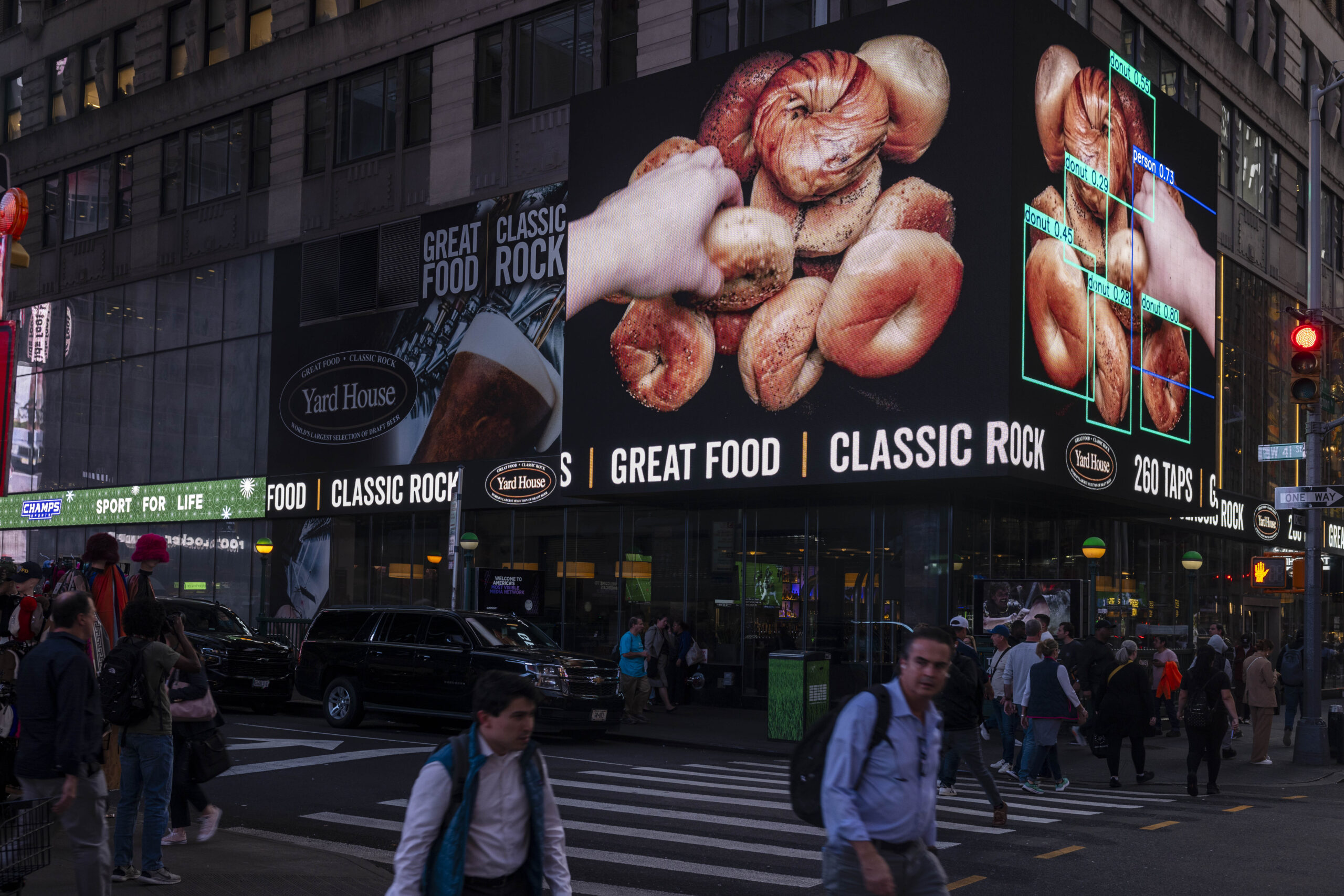 Documentation of a video work created by Ari Temkin. Two large billboard screens wrapped around the corner facade of a building shows a mirrored video of a hand reaching into a pile of bagels. The video on the right has multiple square color marquees of an AI object detection labeling the bagels as donuts.